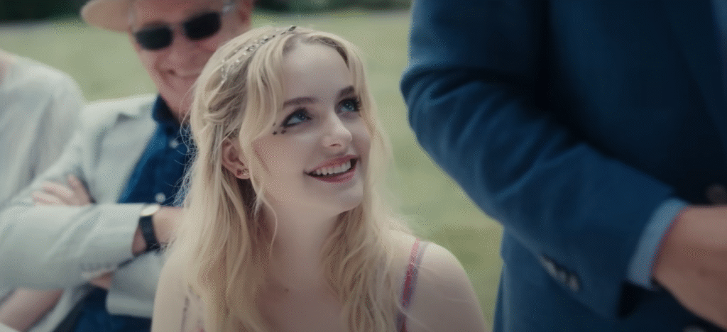 Scene from Anniversary movie 2025 showing a young blonde woman smiling brightly with small star decorations near her eye, dressed in a light outfit, while sitting outdoors during a family gathering. The Anniversary movie features Diane Lane, Phoebe Dynevor, and an ensemble cast, bringing suspense and drama to the big screen in 2025.