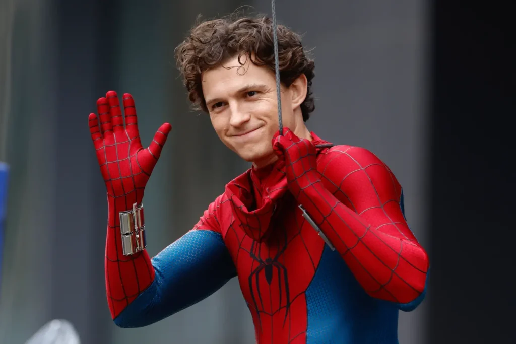 Tom Holland in full costume waving on set during the open shoot of Spider-Man Brand New Day, wearing the new red-and-blue suit while suspended by a wire, captured in a public filming location outside New York.
