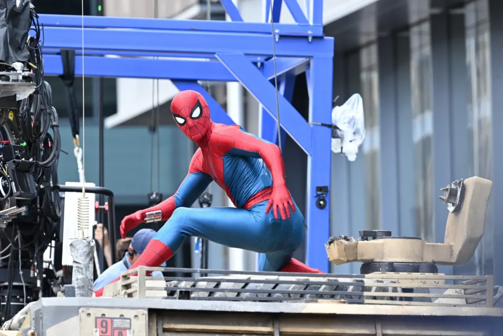 Tom Holland in full costume waving on set during the open shoot of Spider-Man Brand New Day, wearing the new red-and-blue suit while suspended by a wire, captured in a public filming location outside New York.