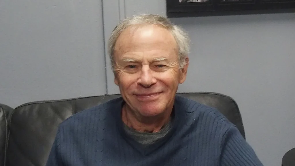 Tristan Rogers smiling in a candid portrait, wearing a dark sweater while seated on a leather chair. The veteran Australian actor, best known for his role as Robert Scorpio on General Hospital, is shown in his later years with gray hair and a warm expression.